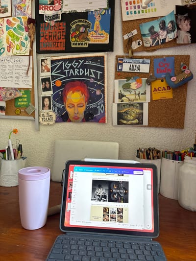 The image captures a vibrant and personalized workspace, likely a desk area, viewed from above. The scene is dominated by a busy cork board on a white wall, filled with a eclectic mix of personal items, notes, and artwork, and a functional work setup on a wooden desk.
On the cork board, numerous items are pinned. In the upper left, there are two photographs: one of a baby and another of three young women. Below these, a small watercolor palette displays color swatches, accompanied by handwritten notes labeled "PALETA DE COLORES" (Color Palette). A small white clip and bobby pins are also secured here. Moving right, a small blue fabric car-shaped object is pinned, along with two fabric patches—one blue with "Cry Baby" in white, and a yellow one with "ACAB" (ALL COPS ARE BASTARD) in black. Below these are a white tag reading "MADRID" and "AMAYA", and two small art prints featuring surreal or fantastical scenes.
In the middle left section of the cork board, a planner or notebook is adorned with stickers, including a vintage "Canadian Pacific AIRLINES" travel sticker, a "HAWAI'I" sticker, an "ABUNDANCE" sticker with a brown animal, and others like "PERROS" (dogs), "CERRITO", and a green sticker stating "MOVEMENT IS MEDICINE". To its right, a prominent poster is displayed, depicting a stylized portrait of a person with orange hair against a cosmic background, featuring text that reads "IGGY TARDUST" and "DOORS OPEN: 1973 SEPTEMBER". Beneath this poster, three small instant-camera-style photos are pinned, one of which includes the text "No Time TO LACE". The bottom left of the cork board contains more personal notes, including a handwritten list titled "METAS 2025" (Goals 2025), surrounded by doodles and a green square sticker proclaiming "ALL POWER TO THE PEOPLE!".
On the wooden desk surface to the right, a white jar brimming with colorful pens and markers, and a wider white container filled with colored pencils are visible. An iPad, housed in a dark grey protective case, is propped on a light-colored stand, connected to a matching dark grey keyboard case. An Apple Pencil is magnetically attached to the side of the iPad. The iPad screen displays a design application, likely Canva, showing multiple pages of a project or presentation. Visible text on the screen includes "HUASTECA", "PROPUESTA VISUAL" (Visual Proposal), and a progress indicator of "27%". Further details like "Página 25 - Agregar título de página" (Page 25 - Add page title) and "Página 26 - Agregar título de página" (Page 26 - Add page title) can be seen. Below the iPad, a light pink cylindrical thermos or mug with a matching lid rests on the desk. Behind it, a small white fluted container serves as a pen holder, containing various pens, including one with an orange flower detail. The scene suggests a creative and productive environment.