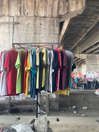 The scene depicts an urban environment beneath a concrete overpass in Aiyetoto-Asogun, Nigeria. In the foreground, a metal clothing rack holds approximately 35-40 short-sleeved T-shirts of various solid colors, including red, light green, yellow, blue, black, grey, white, and maroon, suspended on multi-colored plastic hangers. The rack's central support pole has a black strap wrapped around it, with a small white paper attached. The immediate ground is uneven concrete, strewn with rocks and white plastic debris.

A low concrete wall extends horizontally across the mid-ground. To the right of the clothing rack, an individual wearing a light blue top and dark bottoms is seated on a black motorcycle, facing left. Next to the motorcycle, another individual in a light pink/tan short-sleeved top and dark bottoms stands, also facing left. Several other motorcycles are parked further into the mid-ground.

The background features massive, weathered concrete pillars and the underside of the overpass structure, exhibiting discoloration and visible formwork lines. A lower concrete wall behind the individuals is heavily plastered with numerous torn, faded posters and flyers, partially obscuring a red and orange banner. The ground throughout the visible area is concrete, marked by debris and general wear. No direct interactions between individuals are discernible.