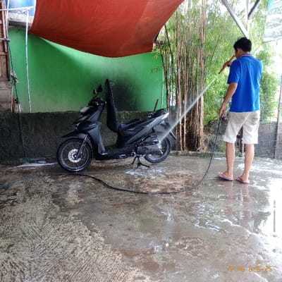 The image shows a man in a blue shirt and black shorts washing a black motorcycle with a hose. The scene is in front of a building with a green wall and a red awning. There are green bamboo plants in the background. The man is holding the hose with both hands and the water is spraying out. The motorcycle is parked on the cement floor. There is a black hose coiled on the ground near the motorcycle. The man is standing on the right side of the motorcycle and the hose is connected to the left side of the motorcycle. The motorcycle is facing away from the man. There is some water on the ground. The location is Kecamatan Legok, Indonesia, based on coordinates (latitude: -6.273, longitude: 106.55).