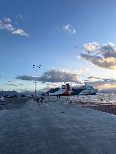 The image depicts a port scene in the city of Aegina, Greece, during late afternoon or early evening under a partially cloudy blue sky. The foreground and midground feature a wide, flat concrete pier with visible expansion joints.

On the pier in the midground, two individuals are riding bicycles away from the viewer; one is on a dark bicycle, and the other on a lighter-colored bicycle. To their left, a line of parked automobiles, predominantly dark-colored, extends into the distance. Several tall, dark streetlights, each equipped with four luminaires, line this section of the pier.

To the right of the pier, two large passenger/vehicle ferries are docked. The prominent ferry displays "saronic" in white lettering on its side, along with the name "PHOIBOS" (ΦΟΙΒΟΣ), and is painted white, dark green, and red-orange. A second ferry, partially visible behind the first, shows its stern in a similar red-orange and white livery. Dark cylindrical fenders are visible in the water near the docked vessels.

Several pedestrians are present on the pier, including two walking near the stern of the main ferry and others further in the distance. A low, orange safety net or barrier, supported by orange cones and poles, lines the edge of the pier adjacent to the water on the right side.

The water of the Saronic Gulf is visible, exhibiting slight ripples. In the background, across the water, hazy blue mountains are visible. The sky transitions from a clear blue overhead to warmer, more illuminated cloud formations near the horizon, consistent with a setting sun.