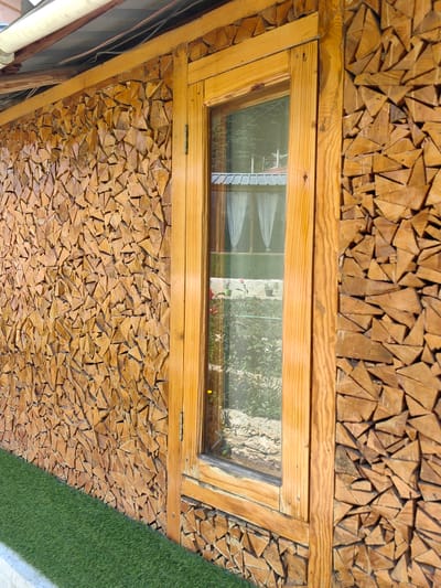 The image shows a wall constructed from small, triangular pieces of wood, resembling firewood neatly stacked and arranged. A wooden-framed window is embedded in this unique wall. Through the window, white curtains are visible, and beyond that, a glimpse of a garden with some plants and soil can be seen. A patch of artificial green turf runs along the base of the wall. The scene is set in Chhyal, India, as indicated by the context. The lighting suggests it is daytime, with sunlight casting shadows and highlighting the texture of the wood. There are no people or animals present in the image. The overall impression is one of rustic architecture and an outdoor setting.