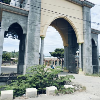A wide exterior shot captures a large, ornate arched gateway, likely the entrance to a mosque or religious complex, in Keudealuerheung, Indonesia. The gateway is constructed from rough-textured grey concrete, with the inner surfaces of the main arch and some upper sections painted a light beige. Its columns feature a speckled, granite-like finish and light green bases.

Through the prominent central arch, a paved courtyard or road leads to other buildings with matching architectural styles, including arched openings and supporting columns. A black metal gate with decorative elements is visible beneath one of the arches on the right. In the distance, beyond these structures, hills covered with lush green trees are visible under a bright, overcast sky. A small dome, typical of mosque architecture, can be seen at the top left, peeking above the gateway's structure.

In the immediate foreground, a vibrant green leafy bush grows beside the road, partially obscuring white concrete blocks lining the edge of what appears to be a drainage ditch. Power lines stretch across the upper portion of the frame, crisscrossing the gateway. A blue barrel and a green, rectangular container are situated in the courtyard beyond the arch. The scene is static, devoid of people or discernible activity, captured during daylight hours under a bright sky. No visible text is present.