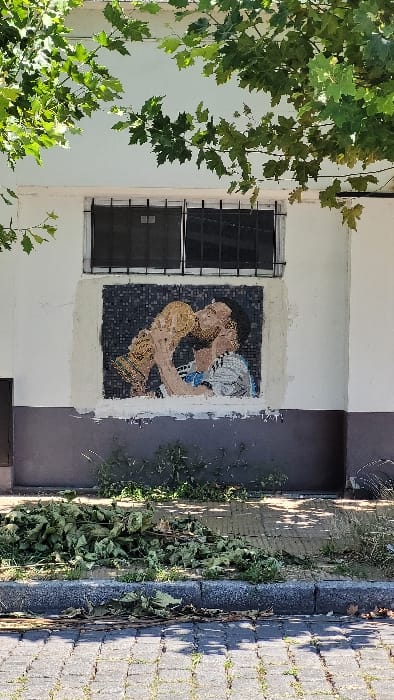 A mosaic art piece depicting two figures embracing is displayed on the exterior wall of a white building. The mosaic is bordered by a lighter shade of color. A barred window is to the left of the mosaic. The building adjoins a dark grey wall that is alongside a sidewalk paved with grey bricks. Green leaves from nearby trees are visible in the upper left corner.