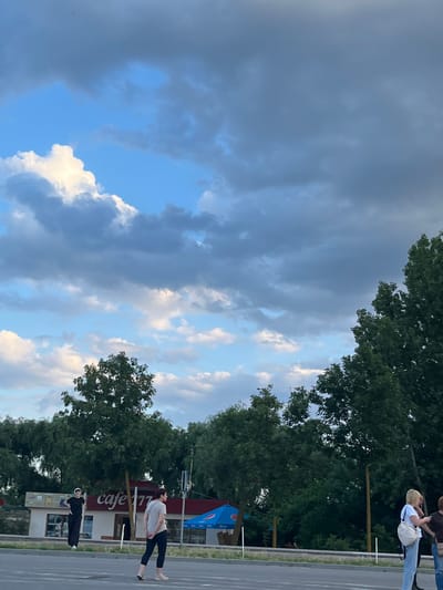 In Palanca, Moldova, an outdoor scene unfolds under a sky filled with dynamic clouds, ranging from bright white cumulus to darker, more imposing formations against a blue backdrop. The lower half of the image is dominated by lush green trees, forming a dense canopy. Beneath the trees, a low-rise building with red accents is visible, featuring the signage "cafe 177". In front of the cafe, a blue umbrella is partially visible. In the foreground, several individuals are present. One person, wearing black attire, stands near the building. Two women are crossing a paved area, with one wearing a light-colored top and dark leggings, and the other in a white t-shirt and blue jeans, carrying a bag. The atmosphere suggests a partly cloudy day with potential for changing weather.