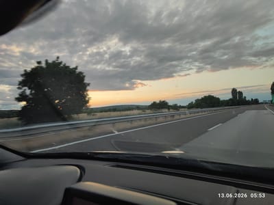 The image depicts a highway scene at sunrise, with a view from inside a vehicle. The sky is filled with dramatic, gray clouds, with streaks of orange and yellow light breaking through at the horizon, indicating the sun is just beginning to rise. A large, dark green tree is prominently positioned on the left side of the road, its silhouette stark against the brightening sky. The highway itself is paved and appears to be relatively empty, with lane markings clearly visible. A metal guardrail separates the road from a grassy embankment lined with more trees in the distance. The foreground shows the interior of the car, including the dashboard and windshield. The timestamp "13.06.2026 05:53" is overlaid on the lower right portion of the image. The scene suggests travel on a highway in a rural or semi-rural area, possibly on the outskirts of Kalugerovo, Bulgaria, given the date stamp. There are no people or active interactions visible in the image.