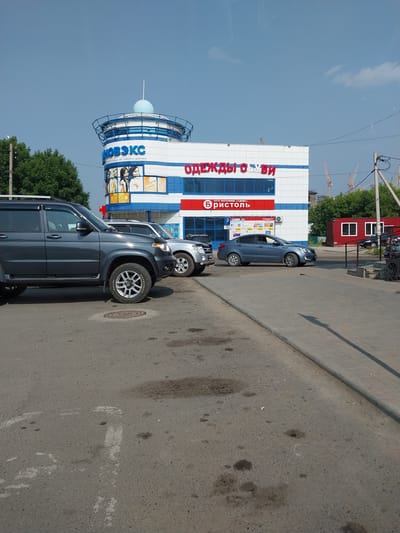 The image shows a street scene in the city of Belokurikha, Russia. The main subject is a white and blue modern building with a rounded top structure. Signs on the building indicate "НОВЭКС" and "ОДЕЖДЫ ОБУВЬ". In front of the building, several vehicles are parked and in motion, including a dark gray SUV, a silver SUV, and a blue sedan. The street is paved and has some visible tire marks and stains. Trees are visible on the left side of the image, and construction cranes can be seen in the background. A red portable structure with windows is also present on the right side of the scene. The sky is clear blue with some faint clouds.