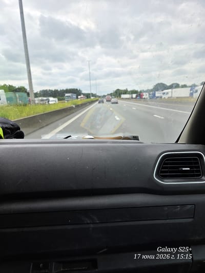 The image displays a view from inside a vehicle, looking out through a slightly dirty windshield. The perspective is from the dashboard, showing the right side air vent and textured dashboard material in the foreground. Beyond the windshield, a highway is visible with multiple lanes and several trucks and cars in traffic. The sky is overcast with clouds. The scene is identified as being in Waasmunster, Belgium, and the date and time stamp "Galaxy S25+ 17 июня 2026 г. 15:15" is overlaid on the lower right corner.