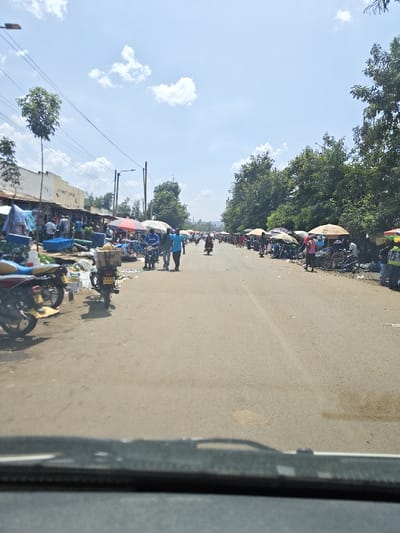 A bustling street scene unfolds with vendors lining both sides, their stalls shaded by umbrellas. People walk and ride motorcycles amidst the market activity. The road stretches into the distance, bordered by trees and buildings. The view is from a vehicle, the dashboard visible at the bottom. The sky is bright with scattered clouds, suggesting a sunny day.