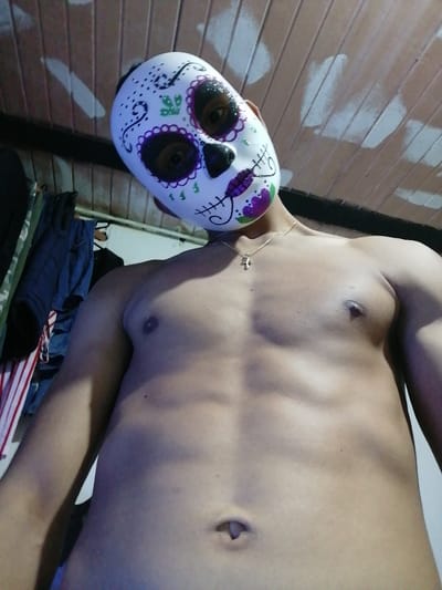 A shirtless person wearing a sugar skull mask poses from a low angle. The mask is white with colorful floral designs around the eyes, nose, and mouth. A thin gold necklace with a cross pendant is visible on the neck. The background shows a slatted ceiling and a closet with hanging clothes. The focus is on the person's torso and mask, creating a unique and slightly unsettling portrait.