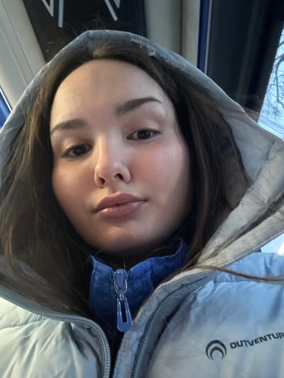 A close-up selfie depicts a young female looking directly at the camera, wearing a light grey-blue puffer jacket with the hood up. Her dark brown hair is visible beneath the hood. She has fair skin, dark eyes, and full lips. A blue garment with a blue zipper and zipper pull is visible underneath her outer jacket. The jacket's right chest area displays a black "OUTVENTURE" logo with a stylized crescent moon symbol. The background is softly blurred, revealing indistinct white shapes on a dark surface to the upper left and bare tree branches against a bright sky through a window on the upper right. The scene is located in the city of Parizhskaya Kommuna, Russia.