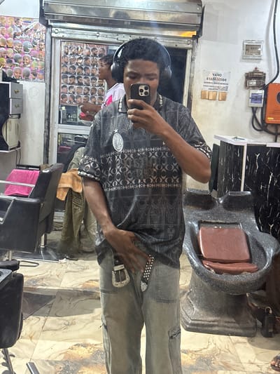 A man is taking a selfie in a barber shop. The location is Ogwashi-Uku, Nigeria. He is wearing a black shirt with a pattern, grey jeans with a studded belt, and headphones. He is holding a smartphone in his left hand. The background of the selfie is the reflection of the barber shop in a mirror. The mirror displays the interior of the shop, including posters of different haircuts and other customers. There is a barber's chair visible to the right. Wiring is exposed near the ceiling.