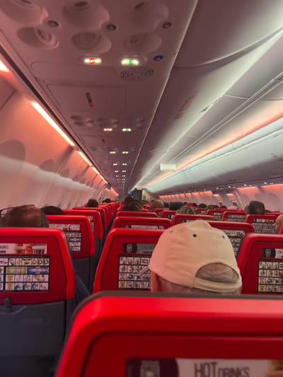 The image shows the interior of an airplane cabin with red seats and orange lighting along the ceiling. The seats have seatback screens displaying various information. Passengers are visible in some of the seats, with their heads and shoulders showing. The aisle of the plane is visible, extending towards the front. The lighting suggests it might be evening or a dimly lit cabin. The plane appears to be on the ground or at a gate, as indicated by the illuminated "EXIT" sign and the presence of passengers. The overall scene depicts a typical commercial airline cabin environment. The location in the city of Lonate Pozzolo, Italy, is a specific detail that cannot be inferred from the image itself, as there are no visual cues to suggest this particular geographical location.