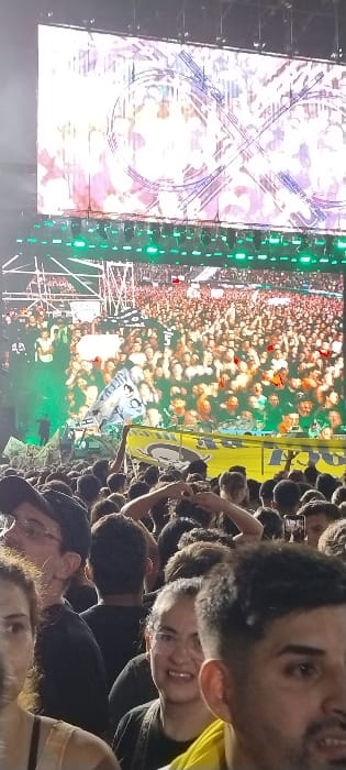 This image captures a vibrant night scene at a large outdoor event, likely a music festival or concert, in Tolosa, Argentina. The photograph is oriented vertically but appears rotated 90 degrees clockwise.
In the foreground, a dense crowd of people is visible. Several individuals are clearly seen, including a man with dark hair and a beard looking towards the camera, wearing a yellow shirt. Beside him, a person with glasses is smiling warmly. Further down, another person with a cap is partially visible. The expressions of those in the foreground suggest enjoyment and engagement with the event.
The mid-ground is filled with an expansive crowd, extending into the distance. Many attendees have their hands raised, and some are holding up flags and banners. A prominent yellow flag with a black design is visible on the right, while a white flag to its left clearly shows the text "AUTIVA" in blue and black lettering. Scattered orange and red elements, possibly confetti or pyrotechnic effects, are seen above the crowd, indicating lively moments during the event. Some people are holding up smartphones, likely capturing the experience.
On the far left, a large, brightly illuminated vertical screen displays abstract, light-colored graphics featuring blue circular and linear patterns. Adjacent to this screen, a tall structure with vertical rows of bright green lights is visible, suggesting part of the stage setup or a lighting rig. The overall lighting, dominated by artificial illumination from the stage and screen, confirms the event is taking place at night. The atmosphere appears energetic and celebratory.