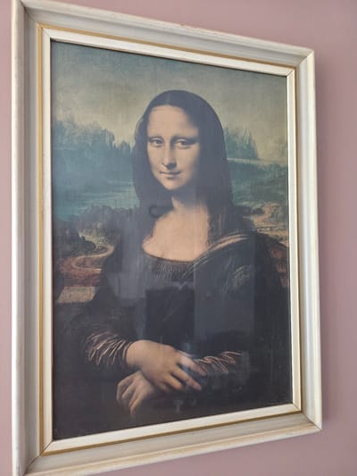 The image is a close-up, angled shot of a framed painting of the Mona Lisa hanging on a pink wall. The painting is a reproduction, evidenced by the slight texture and the visible glare on the surface, suggesting it is under glass. The frame is wide and consists of multiple layers, with an outer layer of distressed light-colored wood and an inner gold-colored accent. The Mona Lisa portrait is centrally positioned within the frame, with her face and upper body visible. Her hands are clasped in front of her. The background of the painting depicts a hazy, mountainous landscape with a winding river or road. The pink wall serves as the immediate background for the framed artwork. The image does not provide any specific information about the city of Brzeg, Poland, or any context related to its location within the city.