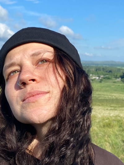 A medium shot captures a woman looking upwards against a bright blue sky with scattered clouds. She is wearing a black beanie and has a silver hoop piercing in her left nostril. Her face is illuminated by sunlight, highlighting freckles across her nose and cheeks. Her dark, wavy hair frames her face. The background consists of a lush green landscape with some buildings visible in the distance. The scene appears to be outdoors, possibly on a hill overlooking a rural area, with no specific indicators linking it to Andreevka, Russia.