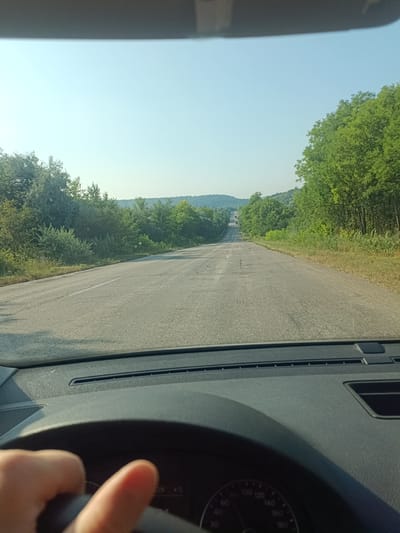 The image depicts a forward-facing view from inside a vehicle traveling on a paved road. The road appears to be cracked and worn, with a white line marking the left lane. Lush green trees and vegetation line both sides of the road, with rolling hills visible in the distance under a clear, pale blue sky. The sun appears to be high, casting a bright light. In the foreground, a portion of the car's dashboard and steering wheel is visible, with a hand holding the steering wheel. The dashboard features a speedometer with markings up to 180 and other indicators. The scene is consistent with a rural or semi-rural environment, typical of the landscape in and around Tibuleuca, Moldova.