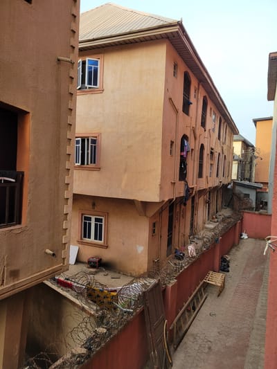 The image depicts an urban residential scene in São Félix do Xingu, Brazil, characterized by closely packed multi-story buildings and a narrow passageway. The primary structure on the right is a multi-story building, likely residential, with a light peach-colored textured facade and a corrugated metal roof. Its exterior features multiple windows, some rectangular with white frames, others with arched tops and fitted with dark metal security bars. Laundry, including shirts and other garments, hangs from several windows and balconies on the upper and middle floors. Exposed utility pipes run vertically along its facade.
To the left, a portion of another building with a darker, reddish-brown textured facade is visible. This building exhibits a distinct stepped, decorative vertical trim. A single open window with dark security bars and a dark interior is present, along with a wooden pipe-like protrusion extending horizontally from its wall.
Separating the main building from a paved alleyway or courtyard is a rust-red painted wall, topped with multiple strands of rusty barbed wire. Debris and various items are entangled within the barbed wire. Behind this wall, on a lower level courtyard, a red portable generator is visible, along with a white panel leaning against the wall and draped fabrics, one red and one yellow-patterned, possibly laundry.
The paved alleyway features interlocking concrete pavers. A wooden ladder lies flat on the ground, and an upright wooden crate or pallet structure is present. Black bags, resembling refuse, are stacked against the wall. In the background, partially visible between the main buildings, are more structures, some with visible corrugated metal roofs and external wiring. The sky above is a pale, uniform grey, indicating an overcast day. No individuals are visible in the scene.