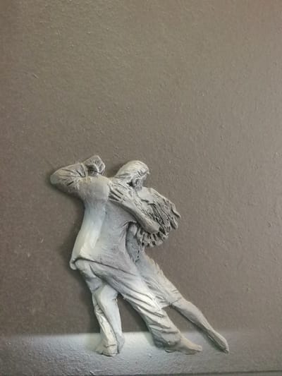 This media file is a close-up, overhead shot of a gray, three-dimensional relief sculpture depicting two people dancing. The figures are stylized and appear to be a couple in a close embrace, possibly engaged in a tango or similar partner dance. The male figure is on the left, wearing what appears to be a suit jacket and trousers, with his left arm raised and bent at the elbow. The female figure is on the right, her body curved and entwined with the male figure. Her hair is depicted as flowing, suggesting movement. The relief is mounted on a textured, dark gray background. The lighting appears to be from the top left, casting shadows that emphasize the contours of the sculpture. There are subtle variations in the gray tones of the relief, suggesting either intentional coloring or the natural effect of light on the material. The bottom edge of the image shows a slightly lighter, perhaps more textured or reflective area, possibly indicating the base or mounting surface of the sculpture. No text is visible in the image.