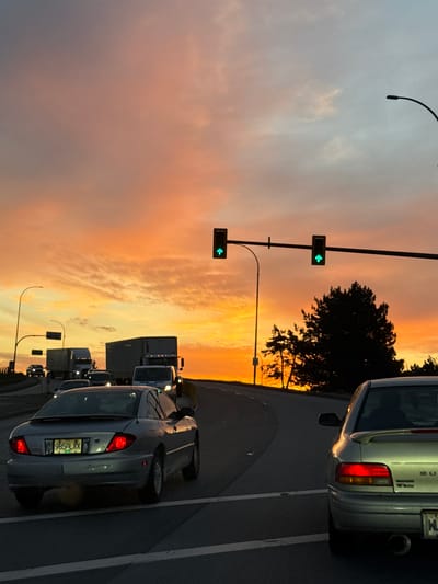 The image captures a street scene in Delta, Canada, during a dramatic sunset or sunrise. The sky dominates the upper portion, displaying a vibrant gradient of warm orange, yellow, and pink hues blending into a softer grey-blue. Diffused clouds are visible across the expanse.

A multi-lane road occupies the lower half of the frame, showing vehicles in traffic, likely stopped or moving slowly. In the immediate foreground, a silver sedan is visible from the rear, its taillights glowing red. Its license plate, a yellow plate with dark characters, reads "965-LXX". Further down the road, a line of semi-trucks and other vehicles extends into the distance, silhouetted against the bright horizon. Another silver car, partially visible on the far right, appears to be a Subaru, with "SUBARU" faintly readable on its trunk lid.

Two traffic lights are prominently positioned on the left side of the road, both displaying green arrows indicating a left turn. Dark, dense trees are silhouetted against the intense sky, adding to the dramatic effect of the low sun. A curved streetlight arm is visible in the upper left, complementing the urban setting. The overall impression is one of a busy roadway during the golden hour, with the striking sky creating a serene yet active atmosphere.