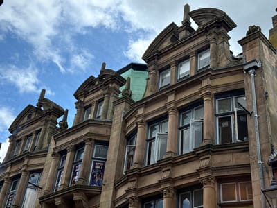 The image shows the ornate facade of a historic building in Leicester, United Kingdom. The building is multi-story with a stone or masonry exterior, featuring architectural details such as columns, arches, and decorative moldings. Several windows are visible, some with advertisements or posters visible inside. The sky is partly cloudy with a mix of blue and gray.