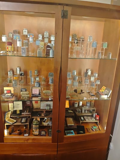 This image depicts an indoor scene in Swindon, United Kingdom, focusing on a tall, wooden display cabinet filled with a collection of vintage lighters and smoking accessories. The cabinet features two hinged glass doors, secured by dark metal latches on the central vertical support. The interior is illuminated, presenting items on four clear glass shelves against a light-brown wooden back panel.
The top two shelves on both the left and right sides contain numerous metallic lighters, primarily silver-toned, but also including gold-toned and brass examples. These lighters are positioned on clear acrylic risers of various heights, creating a multi-tiered display. Observable designs include polished finishes, textured surfaces, and some lighters with colored sections or emblems.
The third shelf from the top, also divided into left and right sections, showcases additional lighters, some contained within their original open presentation boxes. Brands such as "RONSON" are visible on several lighters and packaging, including an open box with an instruction leaflet and a small metal tin labeled "RONSON FLINTS."
The bottom shelf, wider and separated by a horizontal wooden divider, displays a varied assortment. This includes various lighters, a green glass bottle labeled "CLIFTON LIGHTER FLUID," at least one briar tobacco pipe, small wooden display blocks, and several branded boxes such as "ZORRO," "RONSON Varachem Apollo," "EBCO Lighters," and "FIVE LIGHTERS." Other small metal accessories and cases are also present. No individuals are visible within the scene; the items are static and arranged for display.