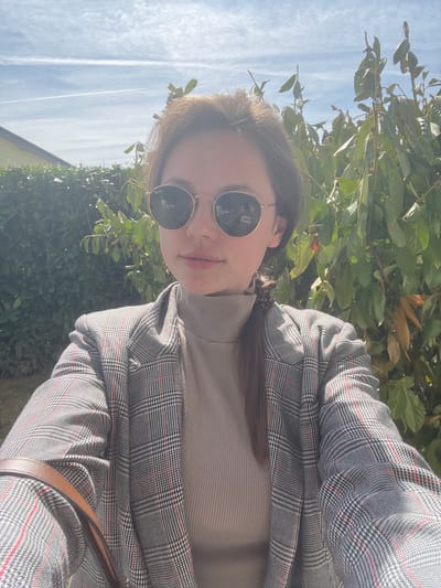 A young woman is posing for a selfie outdoors on a sunny day. She's wearing a light brown turtleneck sweater and a gray plaid blazer. She's wearing round sunglasses and has her hair pulled back into a braid. The background consists of green foliage, suggesting a park or garden. A blue sky with faint cloud streaks is visible above. Given the location of Kranj, Slovenia, it is conceivable she is near the Kokra River or one of the many green spaces within the city.