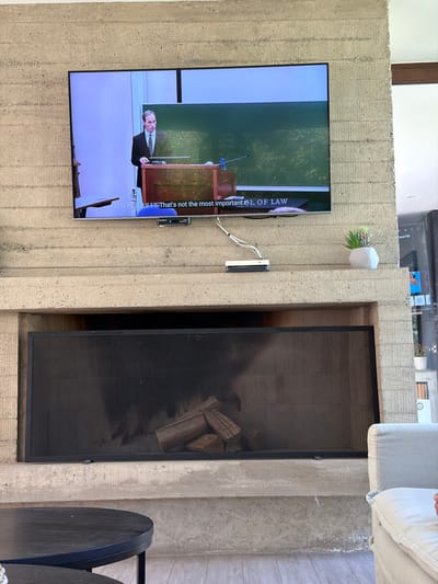 A television shows a person speaking at a podium, a screen reads "That's not the most important dL OF LAW". The TV is mounted to a textured concrete wall, along with a small white object with greenery and a black device. To the right is a fireplace with a black mesh screen and wood inside. In the lower right corner are two black side tables.