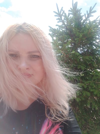 A close-up, outdoor, waist-up shot shows a young woman with blonde hair and a dark shirt with a galaxy-like print. She is outdoors, with a blurry pine tree and a bright sky behind her. Her hair is blown across her face, obscuring part of her features. Her eyes are looking directly at the camera. The lighting suggests it is a sunny day. There is no visible indication of the specific location in Изыхские Копи, Russia.