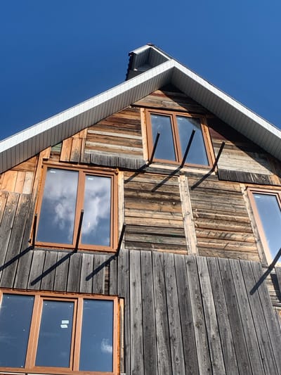 The image displays the exterior facade of a wooden building in Yasinia, Ukraine, viewed from a low angle looking upwards. The structure features a prominent gabled roof with white, horizontally ribbed soffit panels. The building's walls are clad with wooden planks, exhibiting variations in color and texture, from weathered dark grey vertical planks at the bottom to a mix of lighter brown and more aged horizontal planks in the upper sections. Three rectangular windows with light brown frames are visible: two upper windows and one partially visible lower window. The glass panes reflect a bright blue sky with scattered white clouds. Several dark, slender metal bars or supports protrude diagonally downwards from the facade, positioned below the upper windows, casting distinct, elongated shadows onto the wooden siding, indicative of strong, direct sunlight. No people are present.