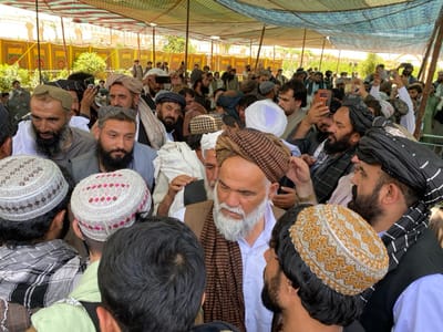 A large crowd of men, mostly wearing traditional Afghan clothing, gather under a white canopy in the city of Zaker-E Sharif, Afghanistan, located at coordinates 31.547, 65.761. Some men are wearing turbans or skullcaps, while others are wearing hats. Many of the men are talking or interacting with each other.  The scene is bright and appears to be taking place during the day. Some men are holding up cell phones, possibly taking photos. Others are holding up cameras, possibly taking videos. The men appear to be gathered for a social gathering.