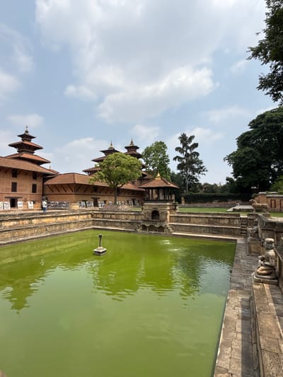 The image shows a pond beside a building in the city of Lalitpur, Nepal. The pond contains green water. A carved stone structure sits in the middle of the pond. The building has a brick facade and tiered, sloping red tile roofs. A tree is planted close to the building. The sky is light blue with scattered white clouds. Other trees are visible in the background. Steps line the side of the pond beside the building. Photos are displayed on the building's exterior. A person is sitting on the steps.