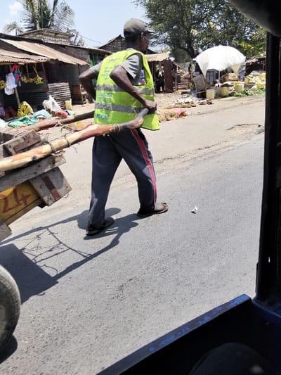 The image captures an outdoor daytime street scene within the Mkomani ward of Kenya. In the midground, a dark-skinned male subject, observed from a rear-right perspective, is actively pulling a wooden, two-wheeled handcart. He is dressed in a grey t-shirt, a neon yellow high-visibility vest prominently displaying "greenlight FINANCING" and two phone numbers ("0725 334 896" and "0725 722 173"), dark grey pants with red side stripes, and dark sandals. A dark baseball cap covers his head. The handcart features a wooden frame and platform, with a yellow numeral "24" visible on its side. The subject is positioned on an asphalt road surface that exhibits minor wear; a small piece of white debris lies on the road near his feet. Strong sunlight is indicated by distinct shadows cast by the man and the cart. The foreground shows parts of a vehicle's blue frame and a tire, suggesting the photo was taken from inside. The background consists of a commercial area featuring multiple makeshift market stalls constructed from corrugated metal sheets and wood, displaying goods such as hanging bananas and yellow buckets. A large white umbrella provides shade over one stall. Accumulations of debris and waste are visible on the ground behind these structures. Palm trees and other sparse vegetation are present under a clear, light blue sky.