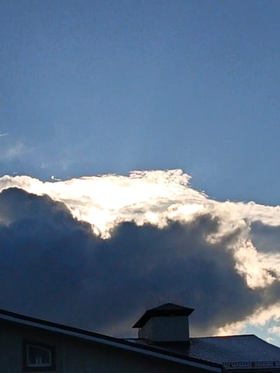 A low-angle outdoor shot captures a dramatic sky above a residential building in Kuyuki, Russia. The upper portion of the image is dominated by a bright blue sky, transitioning to darker hues towards the horizon. A massive cloud formation stretches across the frame, with the top edge brilliantly illuminated by the sun, creating a luminous, almost ethereal glow. The lower portion of the cloud appears dark and dense, suggesting heavy weather.

Beneath the clouds, a portion of a house is visible. It has a dark, sloped roof, likely made of metal tiles, and a light-colored facade. On the roof, a dark, square chimney structure is prominently featured. A single, rectangular window with a white frame is visible on the side of the building. The overall impression is one of dynamic weather conditions observed from a ground-level perspective in a Russian town.
