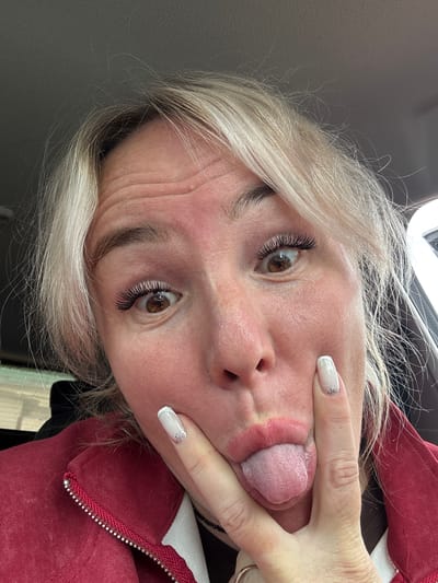 In a close-up, indoor shot, a woman with blonde hair sticks out her tongue and makes a funny face. Her eyes are wide, and her eyelashes are long and dramatic. She's wearing a red jacket, and her fingernails are painted white with glitter. The background is blurry, but it appears to be the interior of a car, suggesting she's on the go. There's no indication of the city of Chastyye, Russia, in this image.