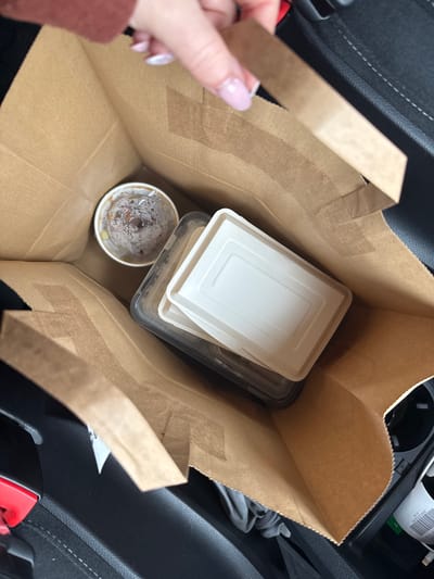 The image shows a brown paper bag filled with food containers, likely takeout. Inside the bag, there are two rectangular containers, one with a white lid and one with a clear lid, stacked on top of each other. Next to them, there is a round container with a clear lid, which appears to have residue suggesting it contained food or liquid. A hand with painted nails is holding one of the bag's handles. The bag is located on the passenger seat of a car, with parts of the car's interior (seat, center console) visible in the background. The location is likely in a car in Amstetten, Austria.