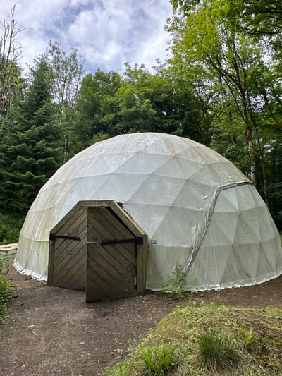 This image shows a geodesic dome structure with a wooden door entrance. The dome is covered in a white material and is surrounded by trees and greenery. The ground in front of the dome is a dirt path. The sky is overcast with clouds. There are no people visible in the image. The scene appears to be in a natural setting, likely a park or wooded area. The image is taken from a slightly low angle, looking up towards the dome and the trees. The overall impression is one of a rustic and somewhat secluded location.