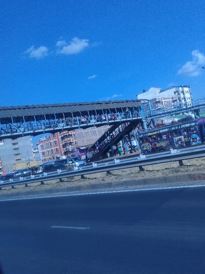 The image captures a vibrant, sunlit urban scene, likely taken from a moving vehicle on a highway. The perspective is slightly tilted, looking upwards and across to the right.

The upper portion of the image is dominated by a clear, bright blue sky with a few scattered, wispy white clouds. The sky appears a uniform, intense blue.

Stretching across the mid-ground is a large, elevated pedestrian bridge, or footbridge, constructed from a blue-painted metal truss structure. The bridge features a covered walkway and multiple sections, including a set of stairs leading up to it. Numerous people are visible on the bridge, indicating a busy thoroughfare.

Below the pedestrian bridge, on what appears to be another road parallel to the foreground highway, several vehicles are present. These primarily look like vans or minibuses. One vehicle, prominently featured beneath the right side of the bridge, is a highly decorated minibus, commonly known as a matatu in East Africa. It is adorned with colorful graffiti artwork, including stylized faces, text, and intricate designs. Visible words on this matatu include "HAWKS" and possibly "CRAZY," though other text is difficult to discern clearly. Other vehicles are parked or moving slowly, suggesting traffic or a pickup point.

In the background, behind the bridge and vehicles, are several multi-story buildings of varying architectural styles. One building on the far right is distinctively white. The overall impression is one of a densely populated urban area.

In the immediate foreground, a dark asphalt highway lane is visible, featuring a faded white solid line. This highway is separated from the activity beneath the bridge by a low concrete barrier topped with a double set of metallic guardrails. The ground between the guardrails and the main road beyond appears dry and dusty. On the concrete barrier in the lower right portion of the image, two red numbers, "8" and "2," are distinctly visible, written as "8+2".

The time of day is bright daylight, indicated by the clear sky and strong illumination. The overall scene conveys the bustling activity of an urban environment, with heavy pedestrian and vehicular traffic. The unique graffiti on the matatu adds a cultural element to the functional infrastructure.