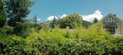 The image shows a view of a building with the text "LA CHOZA" on its facade, set against a backdrop of lush green trees and a clear blue sky with some white clouds. The building appears to be a house or a small establishment with a tiled roof and a prominent chimney. The surrounding environment is heavily vegetated with dense bushes and trees, suggesting a natural or semi-rural setting. The overall impression is one of a tranquil, picturesque location, likely a residential area or a place of leisure. The image does not depict any people or specific actions. The location is described as being in the city of San Rafael, Spain.
