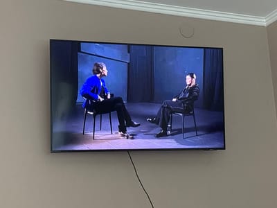 The image shows a television screen displaying two individuals seated on chairs, facing each other. The person on the left wears a bright blue jacket and black pants, while the person on the right is dressed in a black outfit with decorative embellishments on the pants and jacket. Both individuals are looking towards each other, suggesting a conversation or interview is taking place. The background on the screen appears to be a dark, stage-like setting with vertical panels and stage lighting casting shadows on the floor. The television is mounted on a light-colored wall, and a black cable extends from the bottom of the screen. The overall environment is indoors, likely a living room or entertainment area. The television is positioned in the city of Lunnenetsky Sel'sky Sovet, Belarus.
