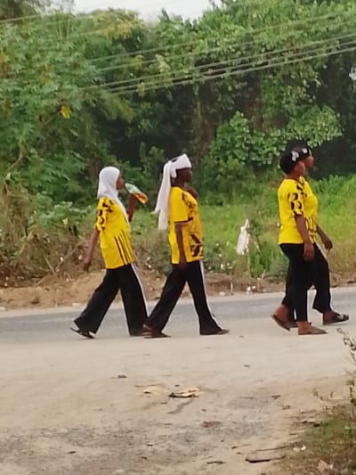 The image captures three individuals, likely women or young adults, walking in a single file line from left to right across an unpaved, dirt road. They are positioned in the mid-ground of the frame.

All three individuals are dressed in matching attire: bright yellow, short-sleeved shirts with black patterns or designs, paired with black pants. The first two individuals from the left appear to have a white stripe running down the outer seam of their black pants. Their footwear consists of simple, open-toed sandals or flip-flops, predominantly in shades of brown or black.

Specific details about each person's appearance:
- The person on the far left wears a white headscarf (hijab) that covers their hair and neck. They are actively drinking from a small, colorful plastic bottle or pouch held up to their mouth. Their yellow shirt features vertical black stripes on the sleeves and possibly on the sides of the torso.
- The middle person has a white cloth or towel draped over their head, tied in a way that allows a portion of it to hang down their back. Their yellow shirt has distinct black geometric patterns on the chest area.
- The person on the far right has dark hair visible, possibly styled, with what appears to be a subtle dark headband or hair covering. Their yellow shirt also displays black patterns on the sleeves and chest.

The activity depicted is a casual walk. Their body postures, with one leg extended forward and the other back, suggest they are mid-stride and moving at a steady pace.

The setting is outdoors, likely in a rural or semi-rural area. The road beneath them is light brown, made of dirt and gravel, with visible small stones and some scattered dry leaves or debris. The background is dominated by lush, dense green foliage, consisting of various trees and bushes, suggesting an environment with abundant vegetation. Thin, dark power lines are faintly visible stretching horizontally across the upper portion of the background, just above the tree line. The lighting appears to be natural daylight, indicating it is daytime, possibly late morning or afternoon, with no harsh shadows, suggesting either an overcast sky or soft sunlight. There are no explicit indicators of weather conditions, but it appears clear. No discernible text is visible on any of the clothing or objects within the image.