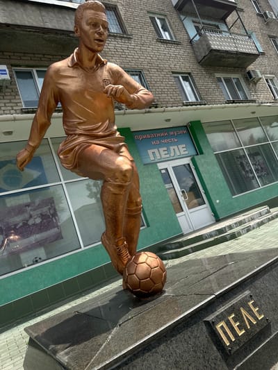This media file shows a bronze statue of a soccer player, likely Pelé, in mid-action, kicking a soccer ball. The statue is in front of a building with a green facade and large windows. Through the windows, reflections of the sky and clouds are visible, suggesting it is daytime with clear weather. The building appears to be a commercial establishment, possibly a museum dedicated to the soccer player, as indicated by the text on a blue sign. The text reads "приватний музей на честь ПЕЛЕ" which translates to "private museum in honor of PELE". To the right of the statue, a dark granite monument bears the name "ПЕЛЕ" in gold lettering. The background shows a brick building with several windows and air conditioning units. The overall scene is outdoors, in what appears to be an urban or semi-urban setting. The statue itself is detailed, capturing the dynamism of a soccer match.