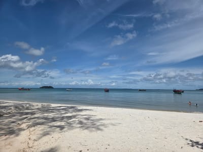 This image depicts a tropical beach scene in Koh Toch Village, Cambodia. The foreground is dominated by a white sandy beach with scattered dried leaves and tree shadows cast across it. The ocean is calm and turquoise, with several boats scattered across its surface. In the distance, a small, tree-covered island is visible. The sky is a vibrant blue with scattered white clouds, some of which appear to be casting shadows on the water. Two individuals are visible in the water on the right side of the frame, appearing to swim or wade. The overall impression is one of a peaceful and idyllic beach day.