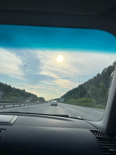 The image shows a view from inside a car on a highway. The sun is visible in the sky, along with clouds. There are trees on the sides of the road and other cars on the highway. The dashboard of the car is also visible in the foreground. The location is the highway near the city of Zvanka, Russia.