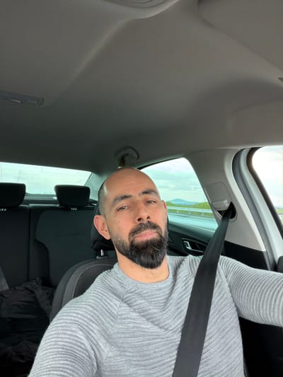 A bald male with a dark beard is depicted taking a selfie inside a vehicle. He wears a light grey, ribbed long-sleeved sweater and is secured by a black seatbelt positioned across his chest and lap. His gaze is directed towards the camera. The vehicle's interior features a light grey headliner. The rear passenger compartment is visible, showing two dark-colored headrests. A small object, possibly a hanger or air freshener, is suspended from the headliner near the driver's side rear door. Through the vehicle's windows, a highway is visible to the right and rear, suggesting movement. Distant hills or mountains under a partly cloudy sky with visible sunlight are present in the background. The scene is located within or in the immediate vicinity of Zetiovo, Bulgaria.