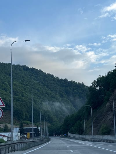 The image shows a highway passing through a mountainous region, likely Surami, Georgia. The road is paved and has white lane markings. On the left side of the road, a steep, densely forested mountain rises, covered in lush green trees. Several lampposts line the side of the highway. In the foreground, a concrete barrier separates the road from the shoulder. On the far left, a traffic sign with a red border is partially visible. In the distance, a tunnel or an opening in the mountain can be seen, with mist or fog surrounding it. The sky is partly cloudy with a soft, diffused light, suggesting it is either early morning or late afternoon. There are no people or vehicles immediately visible in the frame, except for a distant truck.