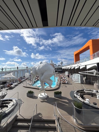The image displays the outdoor upper deck of a modern cruise ship under a clear blue sky with scattered white clouds, suggesting a sunny day at sea. No specific land features of Long Island, Bahamas, are visible; the scene is confined to the vessel.
A prominent, large, white, textured statue of an elephant, appearing to balance on its hind legs atop a smaller, white, quadrupedal creature, is centered on a circular base near an irregularly shaped swimming pool. The pool contains light blue water, and several individuals are present in or around it.
Numerous people are observed on the deck: some are swimming or seated at the pool's edge, while others are relaxing or sunbathing on dark lounge chairs with white cushions arranged around the pool and along the deck. A curving, glass-paneled promenade or upper deck walkway is visible on the left, with several pedestrians walking along it.
Architectural elements of the ship include a distinctive orange, curved structural piece on the right side of the frame, and various white superstructure components, such as radar domes and antennas, are visible in the background. The deck surface is a light brown composite material resembling planks. Additional contemporary outdoor seating areas, complete with curved sofas and small tables, are present on the lower right. Metal-railed staircases lead to lower deck sections, and several planters containing green foliage are strategically placed. The open ocean is visible beyond the ship's railings on the left.