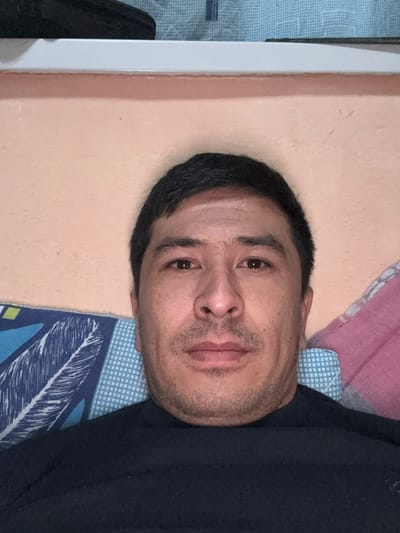 The image displays an adult male, centered in a close-up head-and-shoulders view. He has short, dark hair, brown eyes, a straight nose, and light facial stubble, maintaining a neutral expression while looking directly forward. He is wearing a dark-colored, long-sleeved garment, possibly navy blue or black. The man appears to be reclining, with his head resting on pillows. To the lower left, a pillow with a blue and white patterned case featuring a feather design is visible. To the lower right, a pillow with a light pink or purple patterned case is present. The background includes a pale peach-colored wall, above which a white horizontal surface, likely a shelf or headboard, is seen. At the top edge of the frame, a light blue patterned fabric, possibly a curtain or blanket, is partially visible. The scene is illuminated by artificial light, indicating an indoor setting in the city of Baygundi, Uzbekistan.