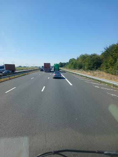 The image shows a view from a vehicle traveling on a multi-lane highway. The sky is clear and blue, and the sun is shining, indicating it is daytime. On the right side of the highway, there is a dense line of trees. On the left side, there is a field with some power lines and distant structures.

The highway appears to be experiencing traffic congestion, with several vehicles visible, including trucks, cars, and a large green vehicle that resembles a tanker or a waste management truck. The vehicles are traveling in the same direction. The road markings, including white lane lines, are clearly visible.

The image is taken from a low angle, suggesting the perspective of a driver or passenger in a vehicle. The foreground shows the dashboard or windshield of the vehicle, with some smudging or reflections.

The specific location is identified as Estrées-Deniécourt, France. This information suggests the highway is likely an autoroute or national road in that region. The presence of power lines and agricultural fields in the background is consistent with a rural or semi-rural setting.