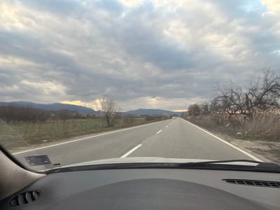 The scene depicts a straight, two-lane asphalt road viewed from inside a vehicle in Novi Chiflik, Bulgaria. The road features solid white shoulder lines and a dashed white center line, extending into the distant horizon. Overhead, the sky is heavily overcast with stratocumulus clouds, pierced by bright light patches and crepuscular rays descending on the right. In the background, mountains with visible snow caps are present. The left side of the road borders agricultural fields containing bare deciduous trees and sparse green ground cover. The right side features an embankment with dry, tall reedy vegetation and leafless trees. Multiple vehicles are observed traveling in the opposing lane in the far distance. The foreground reveals the vehicle's dark dashboard, the lower portion of the windshield, including a reflected sticker on the glass, and a circular air vent on the dashboard's left side.