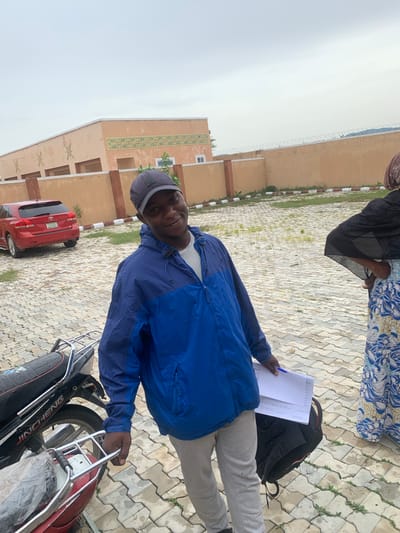 In Wai A, Nigeria, a young man wearing a blue jacket and a grey baseball cap stands smiling, holding a few papers and a pen. He is in front of a red car and a motorcycle parked on a paved courtyard. A woman in a black and blue headscarf and a patterned blue dress is partially visible to the right of the frame. Behind them, a building with a light orange facade and decorative patterns stands tall, enclosed by a light orange wall topped with barbed wire. The sky above is overcast.